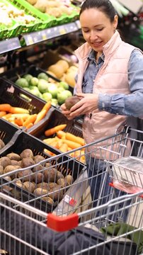 A smiling Caucasian woman selects fresh beets from the counter at a local supermarket and places them in her basket. 4K slow motion footage.