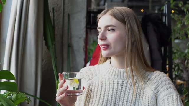 young beautiful woman drinks water with lemon and looks out the window in cafe, girl holds glass and drinks fresh water in restaurant
