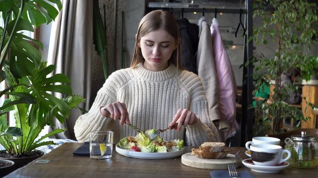 young beautiful woman mixes Caesar salad in cafe, girl with knife and fork eats salad in restaurant