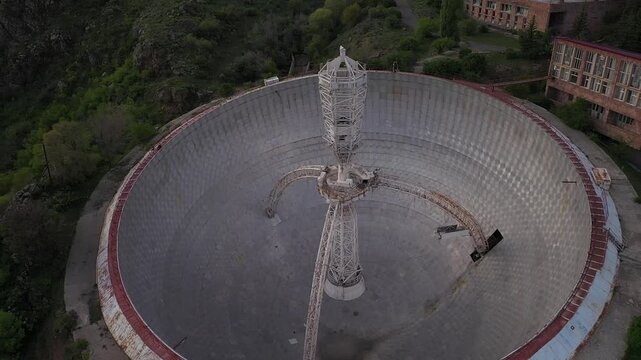  Aerial drone footage slowly zooms in on a massive abandoned radio telescope dish nestled within a mountainous landscape, revealing its intricate central structure and several nearby support buildings