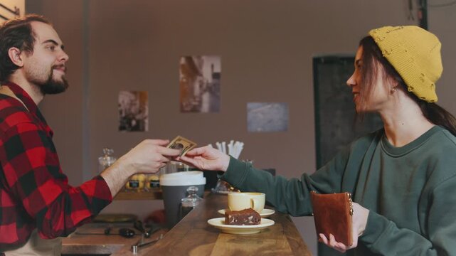 A barista hands a coffee and cake to a customer who gives cash in return. The cafe has a warm atmosphere, with baked goods displayed on the counter.