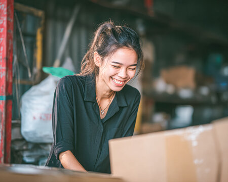 A smiling woman works happily in a warehouse, surrounded by boxes and packaging materials.