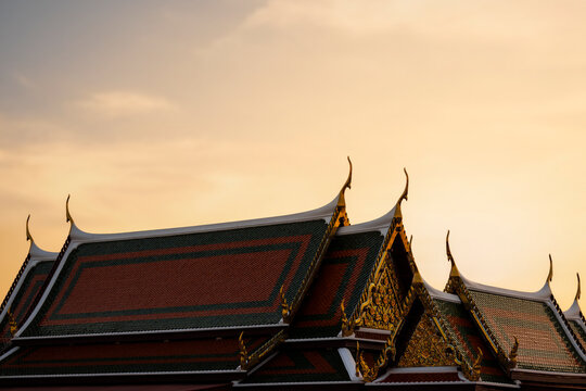 Traditional Thai temple roof with golden chofas and colorful tiles against a warm sunset sky in Bangkok, Thailand.