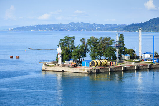 White lighthouse and industrial equipment on Batumi harbor pier, Georgia. Industrial pipes, yellow tanks, and metal tower located behind green trees. Distant hazy mountains visible under sunny sky