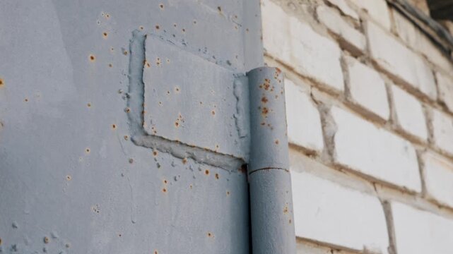 An old rusty metal door hinge attached to a weathered white brick wall. The grey painted steel surface shows early signs of orange corrosion. Heavy welding marks secure the structure.