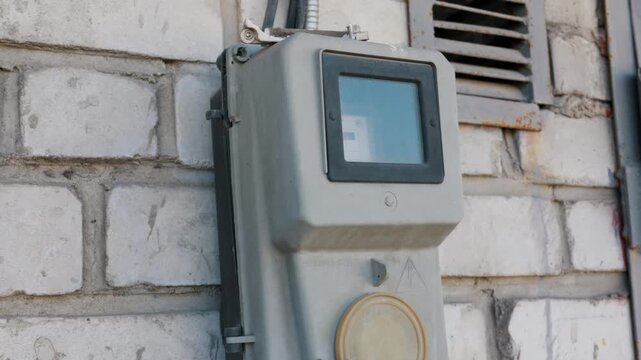 Gray electrical power meter box mounted on an exterior white brick wall of a building. The utility device monitors local energy consumption concept for residential infrastructure.