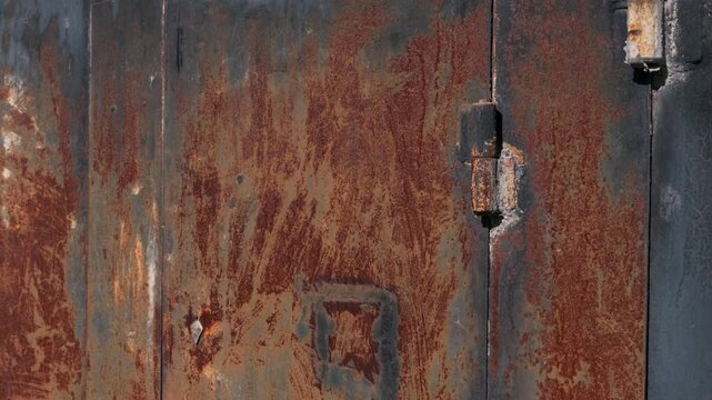 An old rusted metal door stands attached to a white brick wall. Deep orange oxidation covers the rough iron surface. A heavy hinge secures the highly textured industrial structure.
