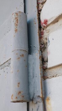 Old rusted metal hinge heavily welded onto a grey painted steel door standing directly beside a textured white brick wall. Concept of industrial aging, structural repair, and urban decay.