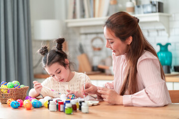 Naklejka premium Happy mother enjoys painting Easter eggs with her young daughter. The little daughter focuses on painting colorful eggs during a fun family craft activity in their kitchen.