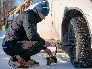 Man inflating car tire with portable compressor outdoors in winter. Driver checking air pressure in wheel on snowy road. Automotive maintenance and vehicle safety service after snowfall.