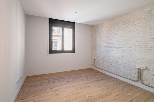 Empty room with wooden floor and white brick wall. A window allows natural light to enter. The space is minimalistic and modern, suitable for various uses.