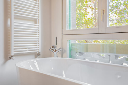 A modern bathroom featuring a white bathtub, a towel radiator, and large windows with a view of greenery outside.