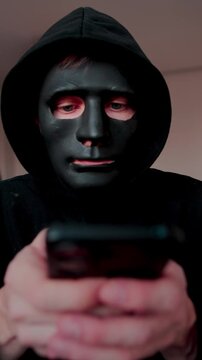 Robber in black mask using smartphone. Hacking, scamming. Vertical video.