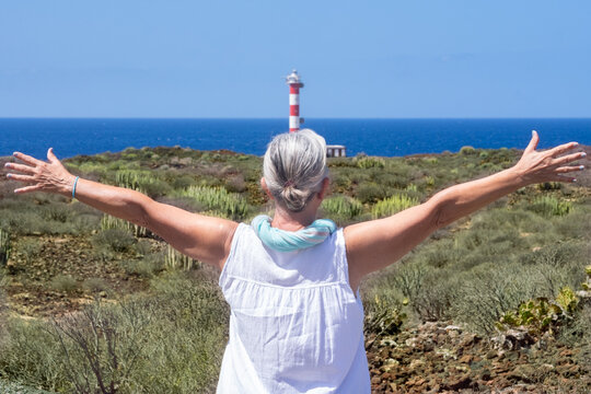 Rear view of a senior woman with outstretched arms enjoying a coastal escursion in a sunny day looking at a red and white lighthouse and the Atlantic Ocean in background.