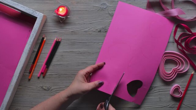 Top view of a hand cutting heart shapes from pink cardboard with scissors. Making Valentine's Day cards, DIY craft and preparation for a romantic celebration