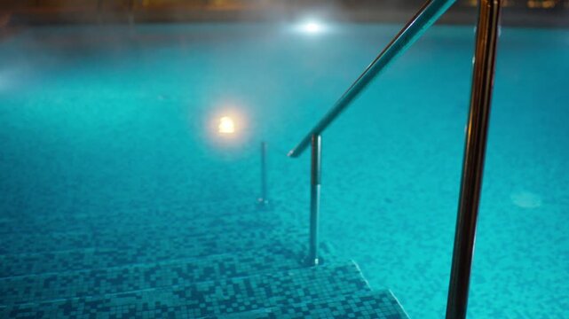 Static shot of a tranquil, illuminated swimming pool at night. Warm, turquoise water steams gently, inviting a relaxing dip down mosaic steps with a gleaming handrail