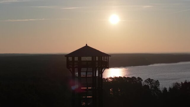 Aerial drone footage of a wooden observation tower surrounded by forest with a lake in the background at sunrise, warm morning light, peaceful nature landscape.