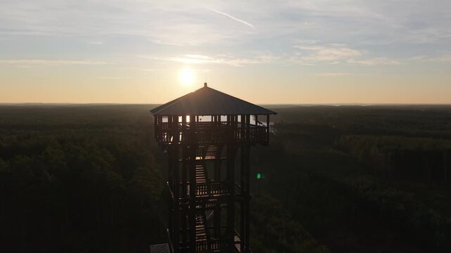 Aerial drone footage of a wooden observation tower surrounded by forest with a lake in the background at sunrise, warm morning light, peaceful nature landscape.