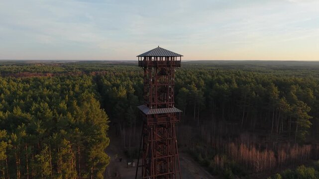 Aerial drone footage of a wooden observation tower surrounded by forest with a lake in the background at sunrise, warm morning light, peaceful nature landscape.