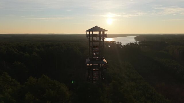 Aerial drone footage of a wooden observation tower surrounded by forest with a lake in the background at sunrise, warm morning light, peaceful nature landscape.
