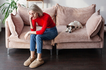 Attractive 40s frustration woman in red blouse with dog sitting on couch indoors. Worried lady have...