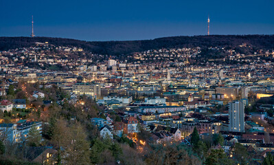 Fototapeta premium Panoramic blue hour view of Stuttgart city center in Germany with glowing city lights, dense urban architecture and residential hills. Television towers visible on the surrounding hills.