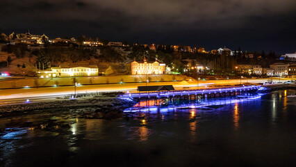 Fototapeta premium Drone view of Akureyri in North Iceland at night with illuminated bridge and city lights reflecting on water. Beautiful aerial scene of Akureyri harbor showing urban landscape and coastline in Iceland