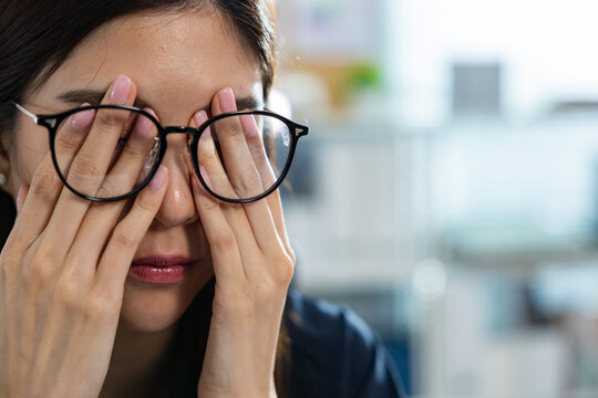 Exhausted Asian woman feeling tired and experiencing deep stress at office rubs her eye under glasses to relieve pain and exhaustion during difficult business day filled with heavy pressure