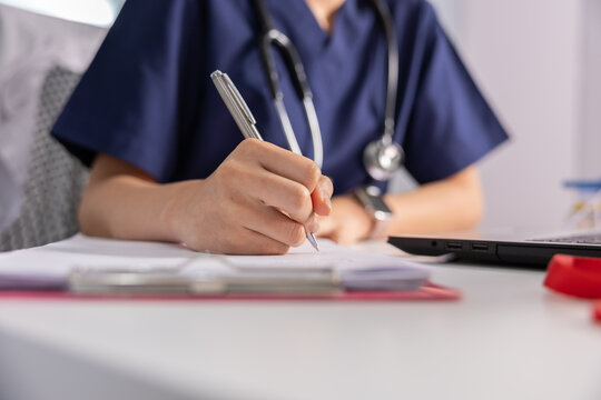 Female doctor nurse in blue scrubs writing notes on clipboard using pen with stethoscope around neck working in hospital clinic showing focused calm emotional expression