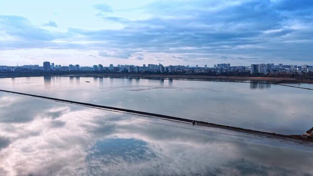 Footage over the vast ponds used for production of salt. Cloudy sky reflects in the waterscape of salterns. City skyline in the backdrop.
