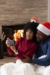 Naklejka premium African American couple sitting by fireplace, taking selfie on phone, Santa hat, snuggling blanket