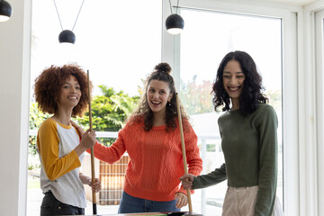 Diverse female friends playing pool at home, holding cues around green felt table near windows