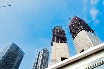 Obraz premium Skyward towers rise against clear blue sky. Construction cranes hover above steel frameworks. Modern architecture dominates urban skyline. Distant skyscrapers reflect sunlight and shadow
