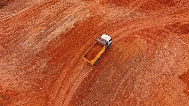Truck loaded by the red soil goes slowly by the rocky area. Arid mountains with poor vegetation in the backdrop. Aerial view.
