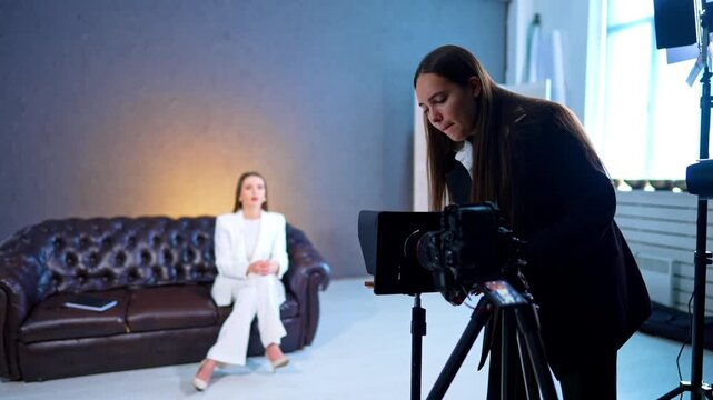 Brunette lady in black suit arranges teleprompter at the modern camera set in studio. Woman in white suit sits on the sofa in the blurred backdrop. Blog content creation.