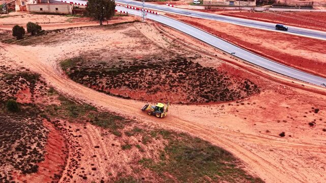 Yellow backhoe loader driving on red soil dirt road. Aerial high angle view of a construction machine on a dusty path. Red earth landscape with rural infrastructure.