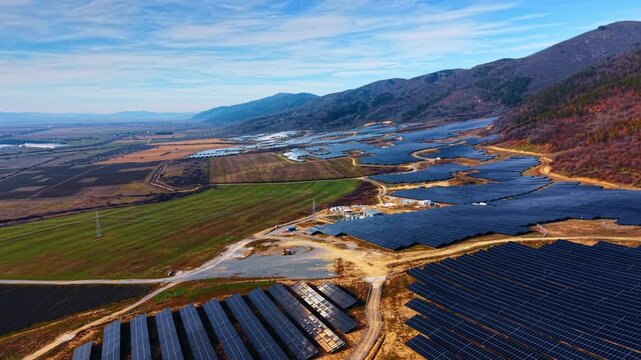 Solar energy farm installation on agricultural fields near mountains. Aerial perspective of solar panels rows integrated into rural landscape with green fields and distant mountain peaks.