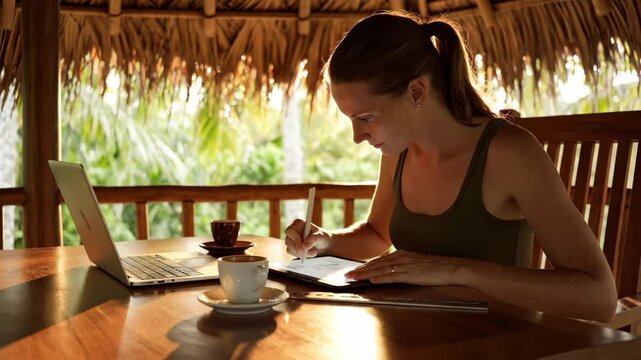 Woman writes on tablet in tropical hut with laptop and coffee. Freelancer works with tablet under thatched roof. Woman uses tablet and laptop in tropical workspace. Coffee beside working woman.
