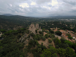 Obraz premium Aerial view of ancient stone castle, Chateau de Grimaud ruins atop a verdant, tree covered hill overlooking a French village with terracotta rooftops and a distant coastline under a cloudy sky. 