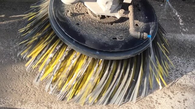 Slow motion close-up of a construction sweeper side brush rotating with high-pressure water jets on a dusty road.