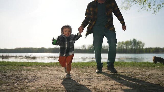 Father, child running hand in hand. Happy child holding father hand, running outdoors. Family bonding moment by the lake. Joyful father and child playing together. Child run with father hand