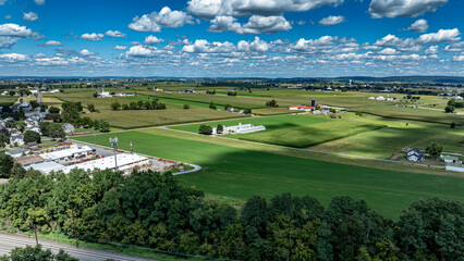 Fototapeta premium The scene shows a broad view of fields and farms located near a small town. Green fields stretch across the landscape under a bright blue sky with scattered clouds. Buildings and trees are visible.