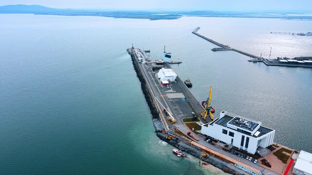 Port infrastructure with modern architecture and coastal work. Aerial view of a port building, parking lot, and dredging boat near shore.