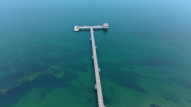 Long wooden pier stretching into calm turquoise sea water. Minimalist top view of a straight T-shaped pier over clear ocean water.