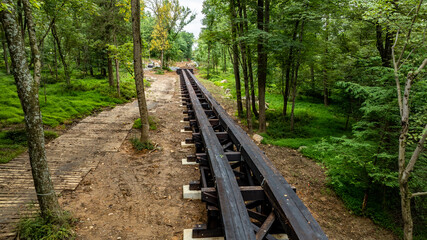 Workers build a wooden railway track in a forest. Trees surround the area. A walking path runs alongside the construction. Tools and equipment are visible in the distance.