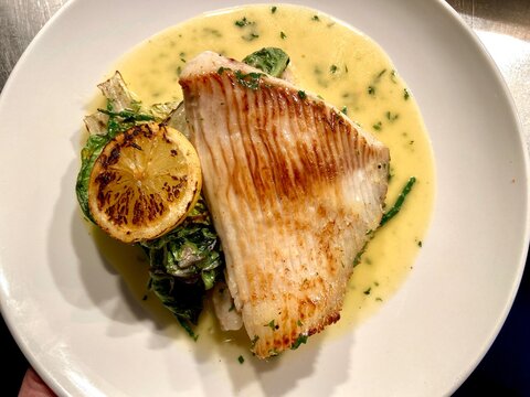 Pan-fried skate wing fillet served with creamy lemon butter sauce, charred lemon, and seasonal greens on a white plate. Elegant seafood dish ideal for restaurant menus, fine dining, British cuisine, 