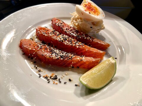 Beautifully presented seared salmon sashimi served with toasted sesame seeds, fresh lime wedge, and a creamy garnish on a white plate. Gourmet seafood dish ideal for Japanese cuisine, fine dining, hea