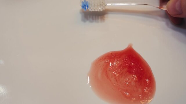 Close-up shot of a bathroom sink during a morning hygiene routine. The person spits into the sink, and the saliva appears slightly bloody, suggesting gum irritation or bleeding during brushing. High