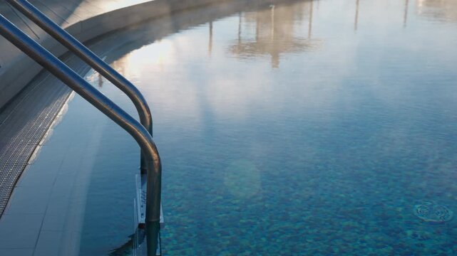 A serene shot of a warm outdoor swimming pool on a cool day, with gentle steam rising from the blue water and sunlight reflecting on the surface
