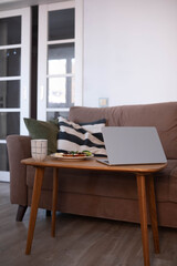 A cozy living room setup: a brown sofa with patterned cushions, a wooden coffee table with a laptop, a plate of fresh snacks, and a white mug with a grid design.